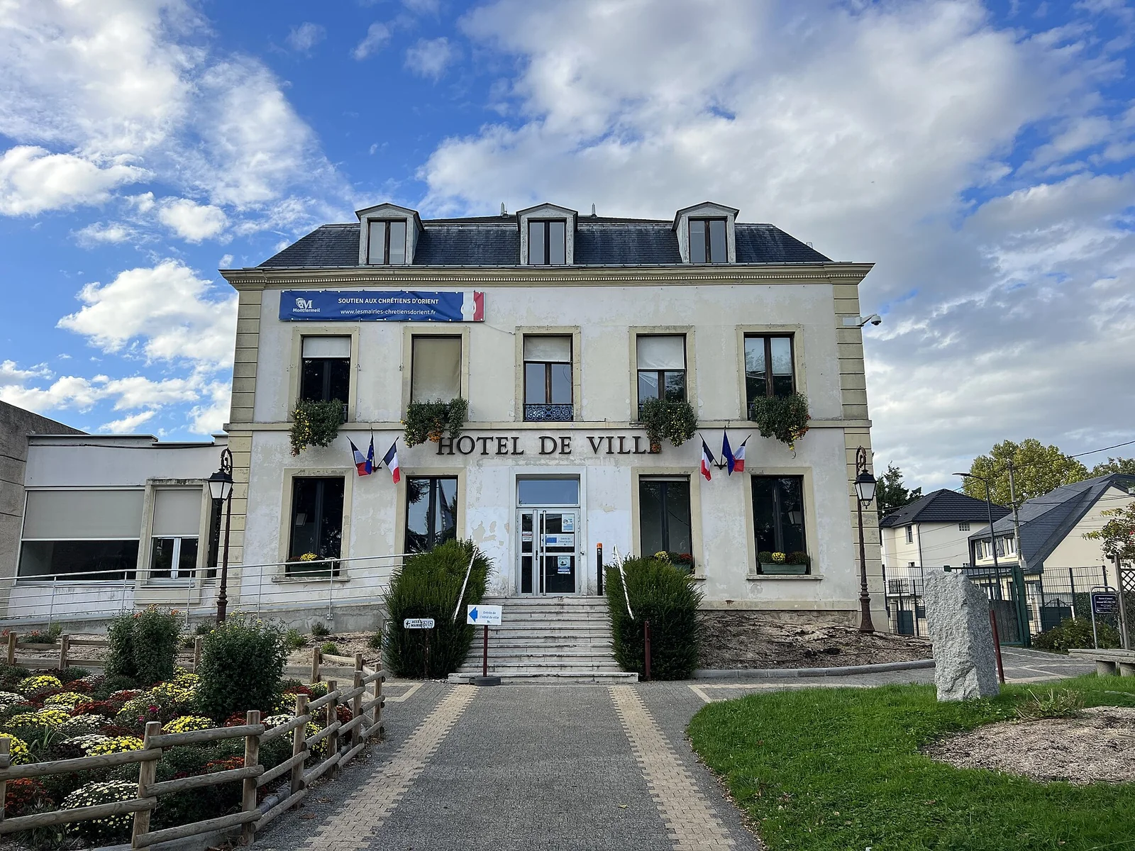 The pale façade of Montfermeil town hall, with French flags, a central entrance and planted beds beneath a cloudy blue sky.