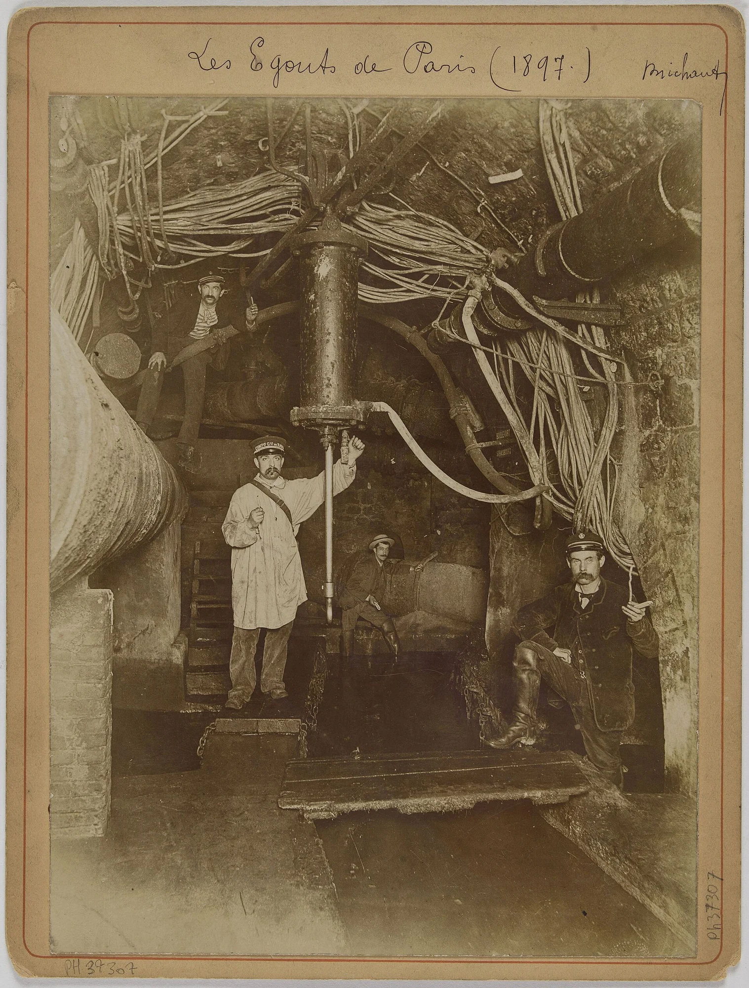 A mounted sepia photograph showing men, pipes and cables inside a Paris sewer tunnel.